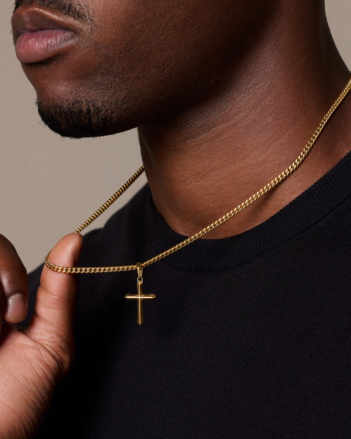 A close-up of a man wearing a gold cross pendant on a gold cuban link chain, paired with a black t-shirt against a neutral beige background.