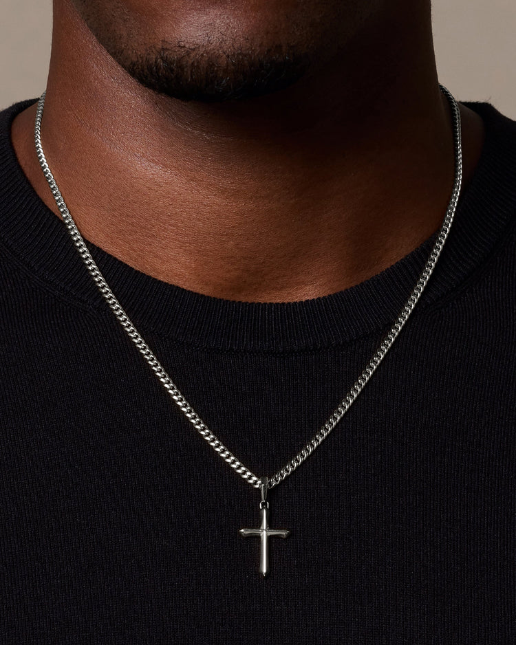 A close-up of a man wearing a silver cuban chain necklace with a small cross pendant, paired with a black crewneck shirt against a neutral beige background.