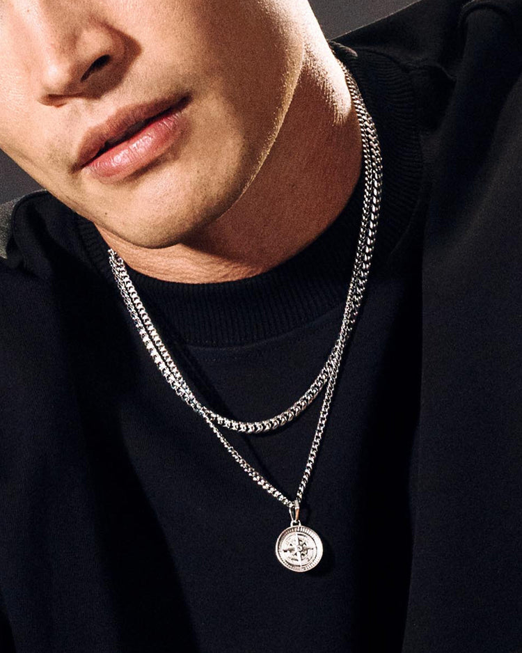 A close-up of a man wearing a double-layered silver chain necklace with a round compass pendant, styled with a black crewneck shirt.