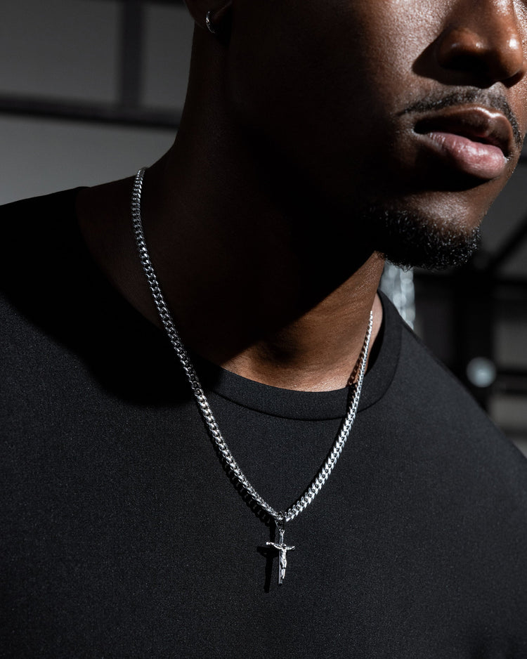 A close-up of a man wearing a silver cuban chain necklace with a small crucifix pendant, styled with a black crew-neck t-shirt.