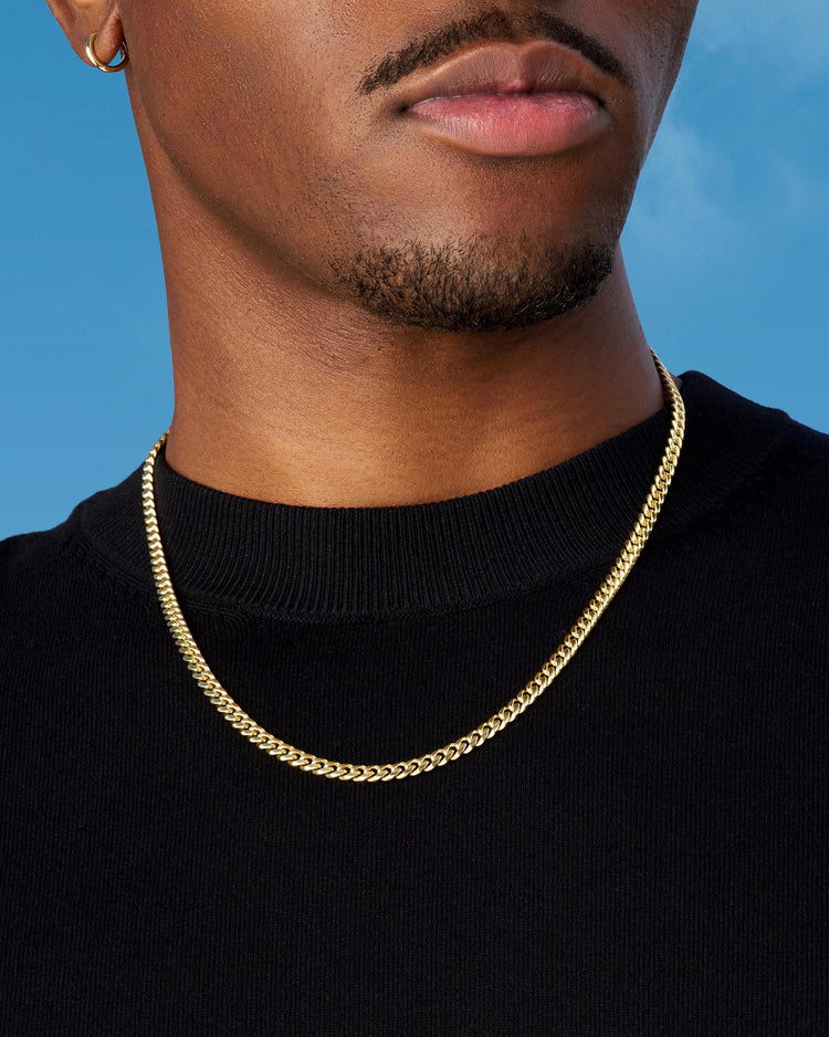 Man wearing gold Cuban chain, black sweater, against a blue sky.