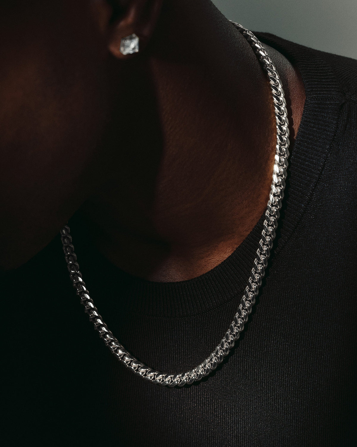 A close-up lifestyle shot of a man wearing a silver Cuban link chain necklace and a matching silver stud earring, wearing a black crewneck shirt.