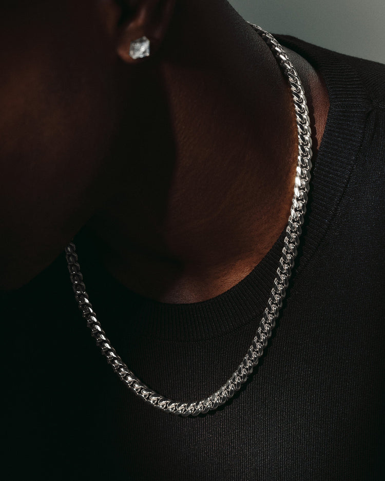 A close-up lifestyle shot of a man wearing a silver Cuban link chain necklace and a matching silver stud earring, wearing a black crewneck shirt.