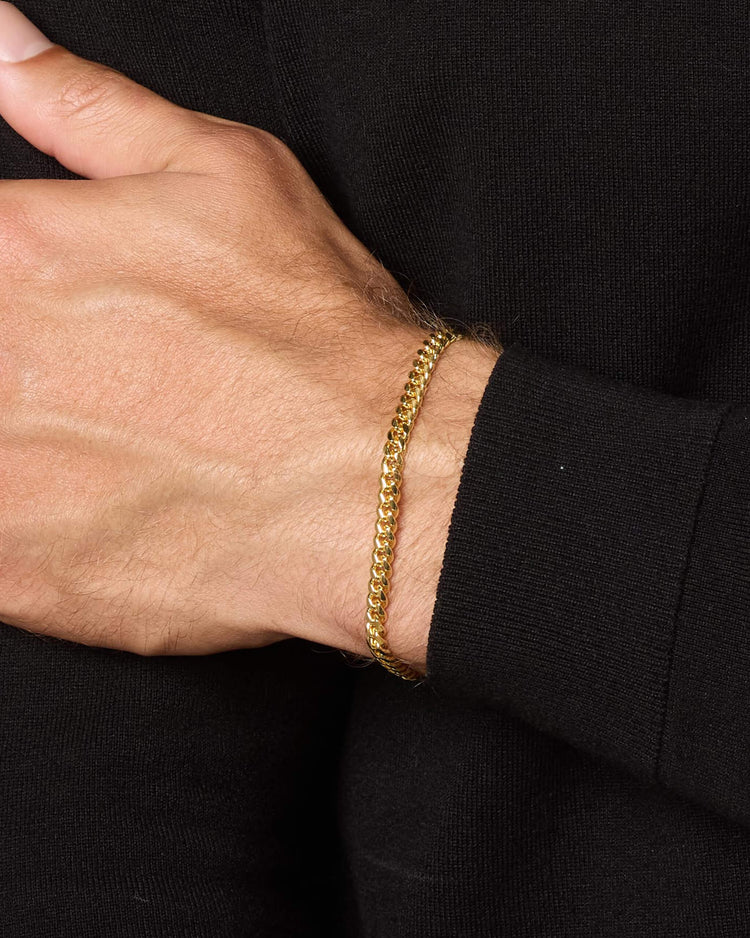 A close-up of a person's wrist wearing a thin yellow gold chain bracelet, shown against a black long-sleeve shirt.