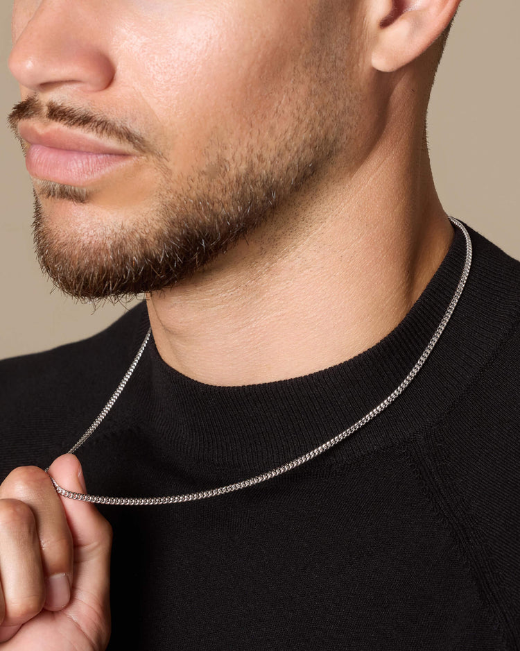 A close-up of a man wearing a thin silver Cuban link chain necklace over a black crewneck sweater, with his hand lightly touching the chain.