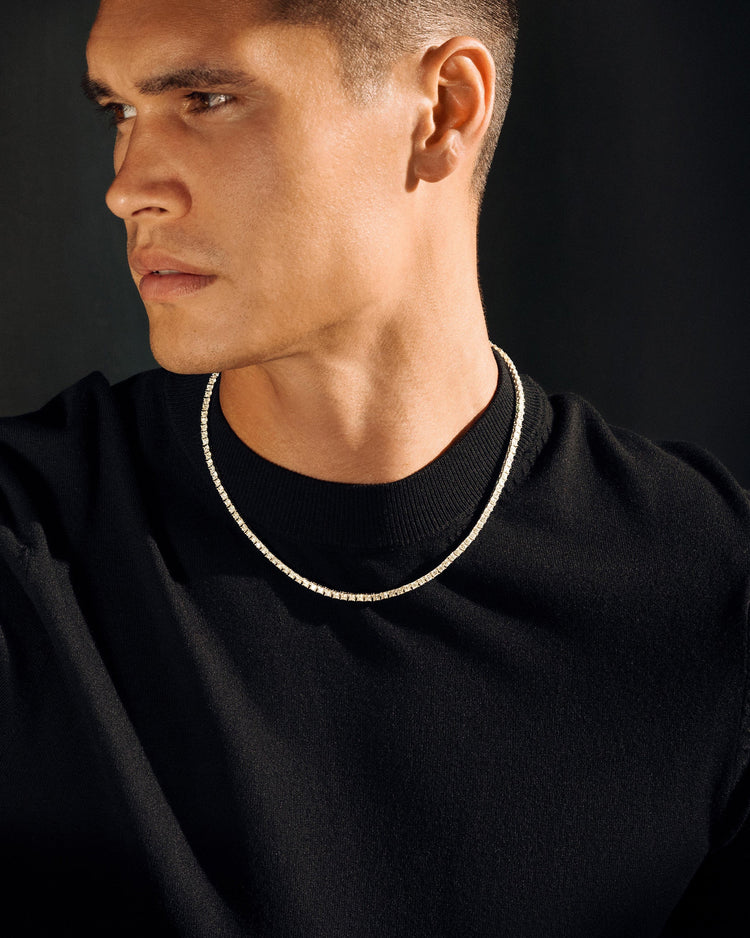 A close-up of a man wearing a thin silver chain necklace over a black crewneck shirt, set against a dark background.