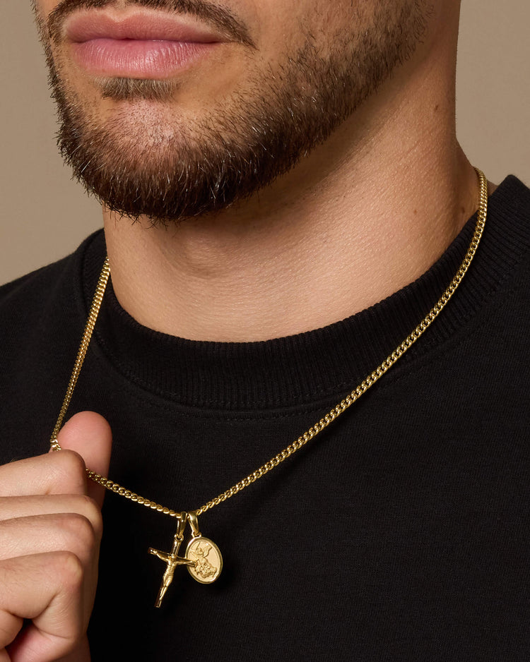 A close-up of a man wearing a gold Cuban chain necklace with a crucifix pendant and a small round St. Michael pendant, holding the chain with one hand against a black crewneck shirt.