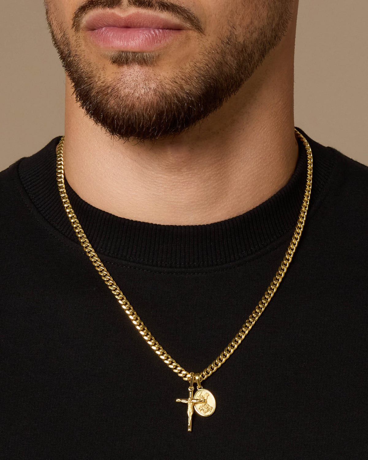 A close-up of a man's neck and lower face wearing a gold Cuban link chain necklace with a Crucifix  and small St. Michael medallion pendant, paired with a black crewneck shirt against a warm beige background.