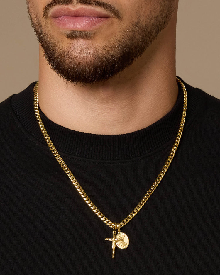 A close-up of a man's neck and lower face wearing a gold Cuban link chain necklace with a Crucifix  and small St. Michael medallion pendant, paired with a black crewneck shirt against a warm beige background.