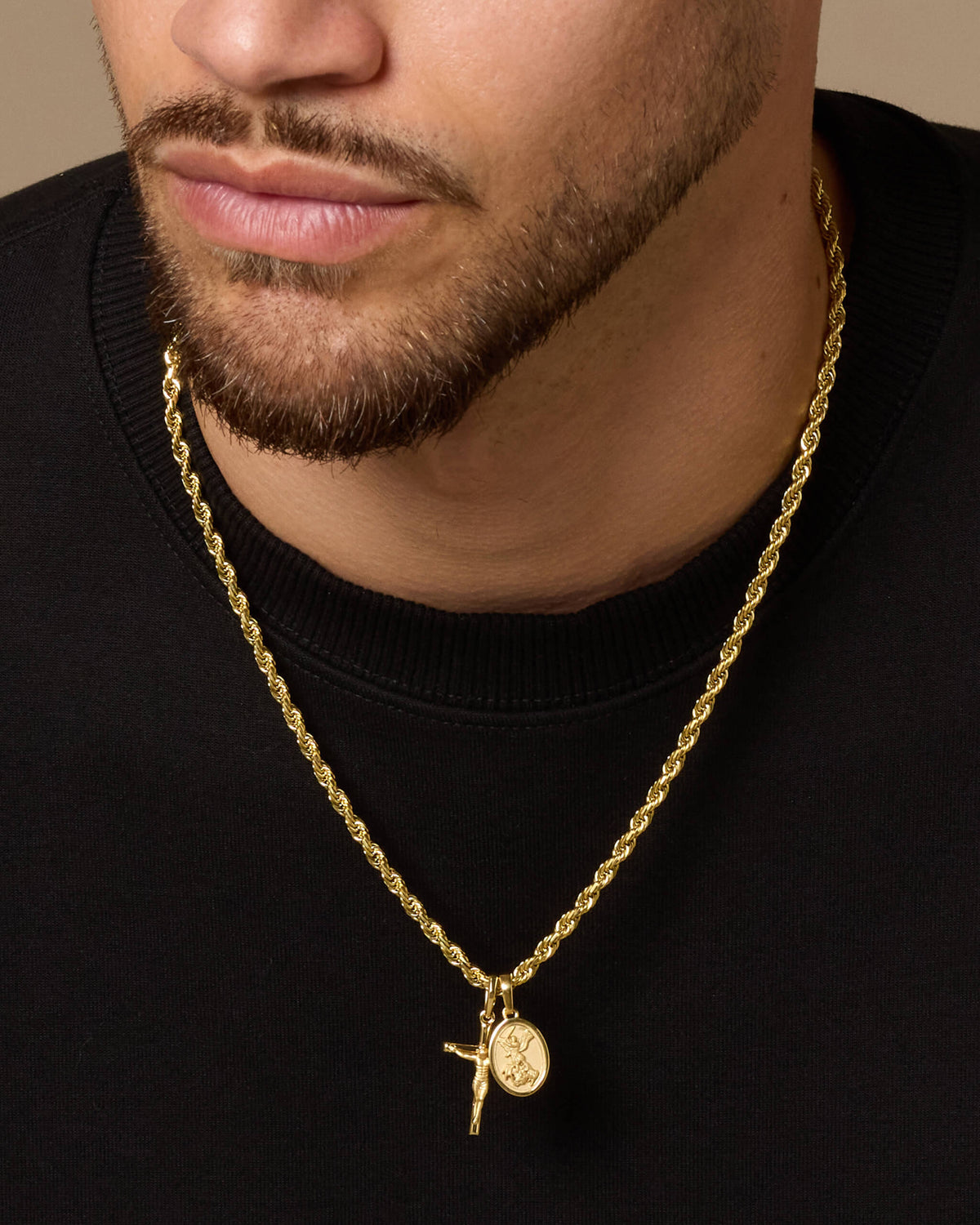 A close-up of a man wearing a gold Rope chain necklace with a crucifix and St. Michael medallion pendant, styled with a black crewneck shirt.